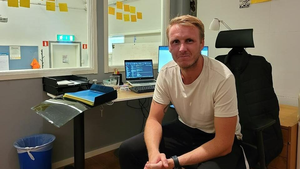 Oscar Lönnerheden sitter på en svart kontorsstol på ett kontor. I bakgrunden syns en dator och flera datorskärmar. Han har vit tröja och svarta byxor. 