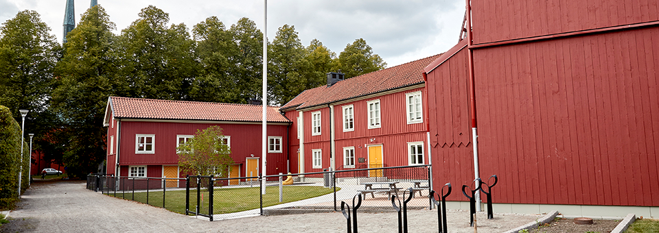 På bilden ser vi ett stort rött trähus med rött tak och gula dörrar. På förskolans innergård finns en liten gräsplätt med en flaggstång på.