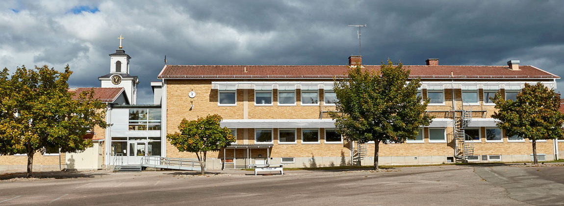 Gul tegelbyggnad med tre gröna träd på skolgården framför skolan