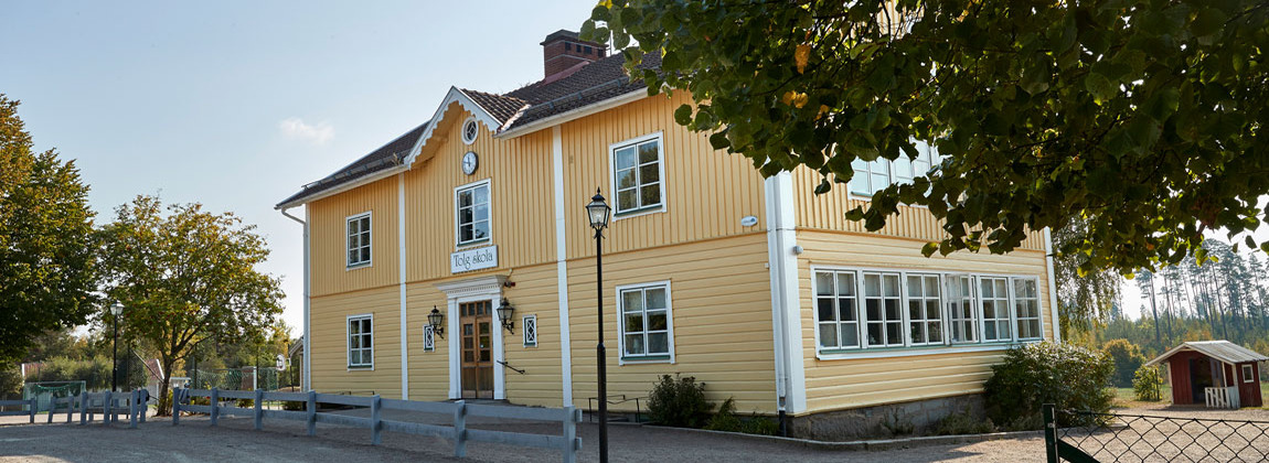 En gul gammaldags träbyggnad med vita fönster. En stor brun trädörr med fönster. Bakom skolan står en röd lekstuga