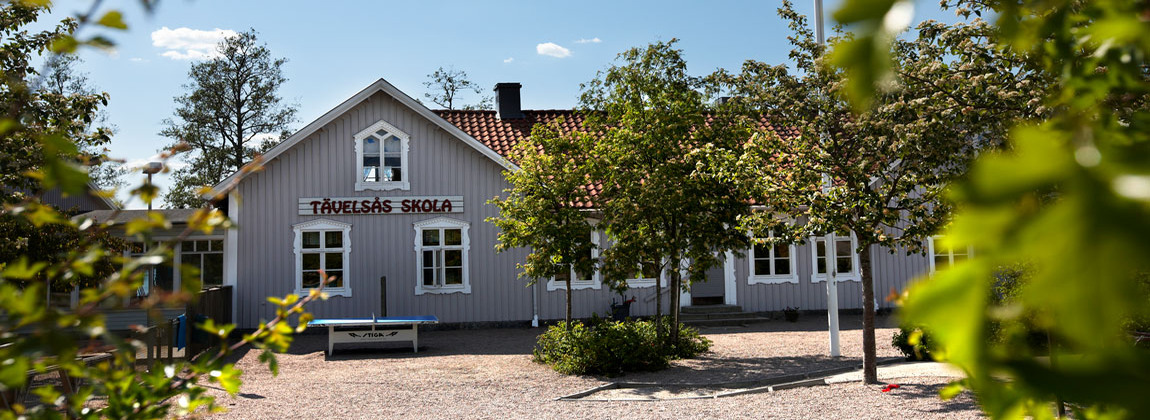 Grå träbyggnad. På byggnaden står det Tävelsås skola i vita bokstäver. Ett pingisbord står på skolgården framför skolan