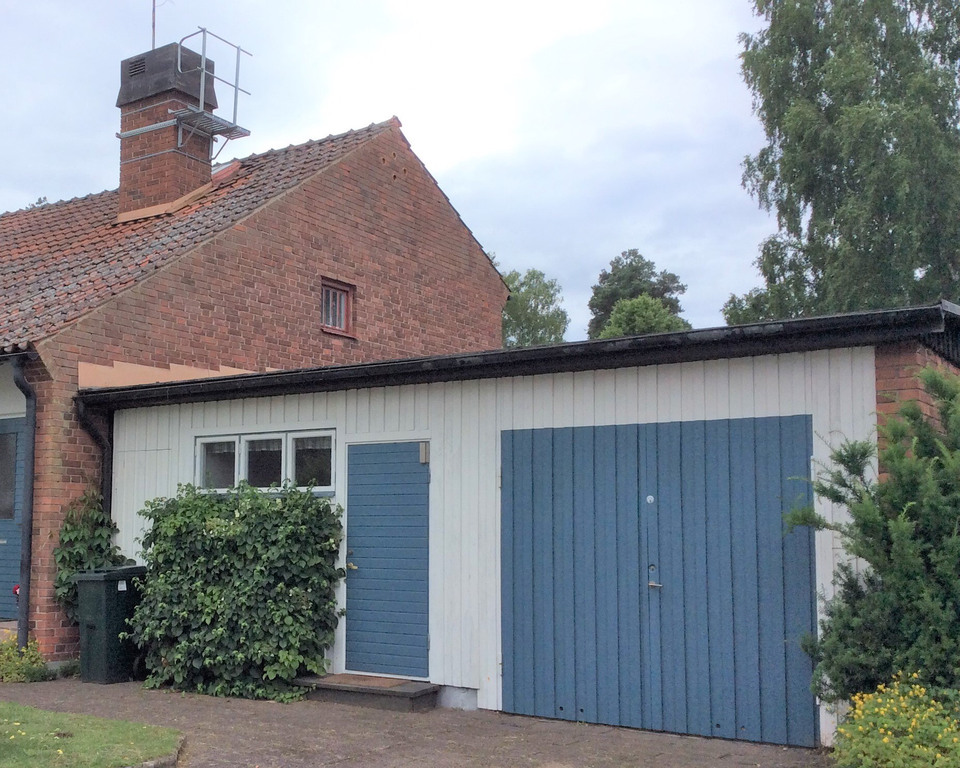 Garage med plant tak i galvad plåt av svart kulör och fasad med vit stående träpanel.