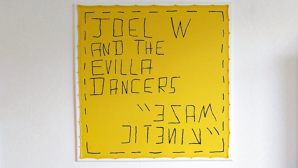 Gult textilverk med svart broderat text: JOEL W AND THE EVILLA DANCERS.